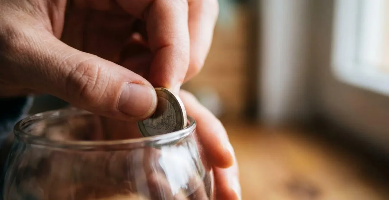Gros plan sur des mains déposant délicatement une pièce dans un contenant d'épargne symbolisant la construction progressive d'une sécurité financière